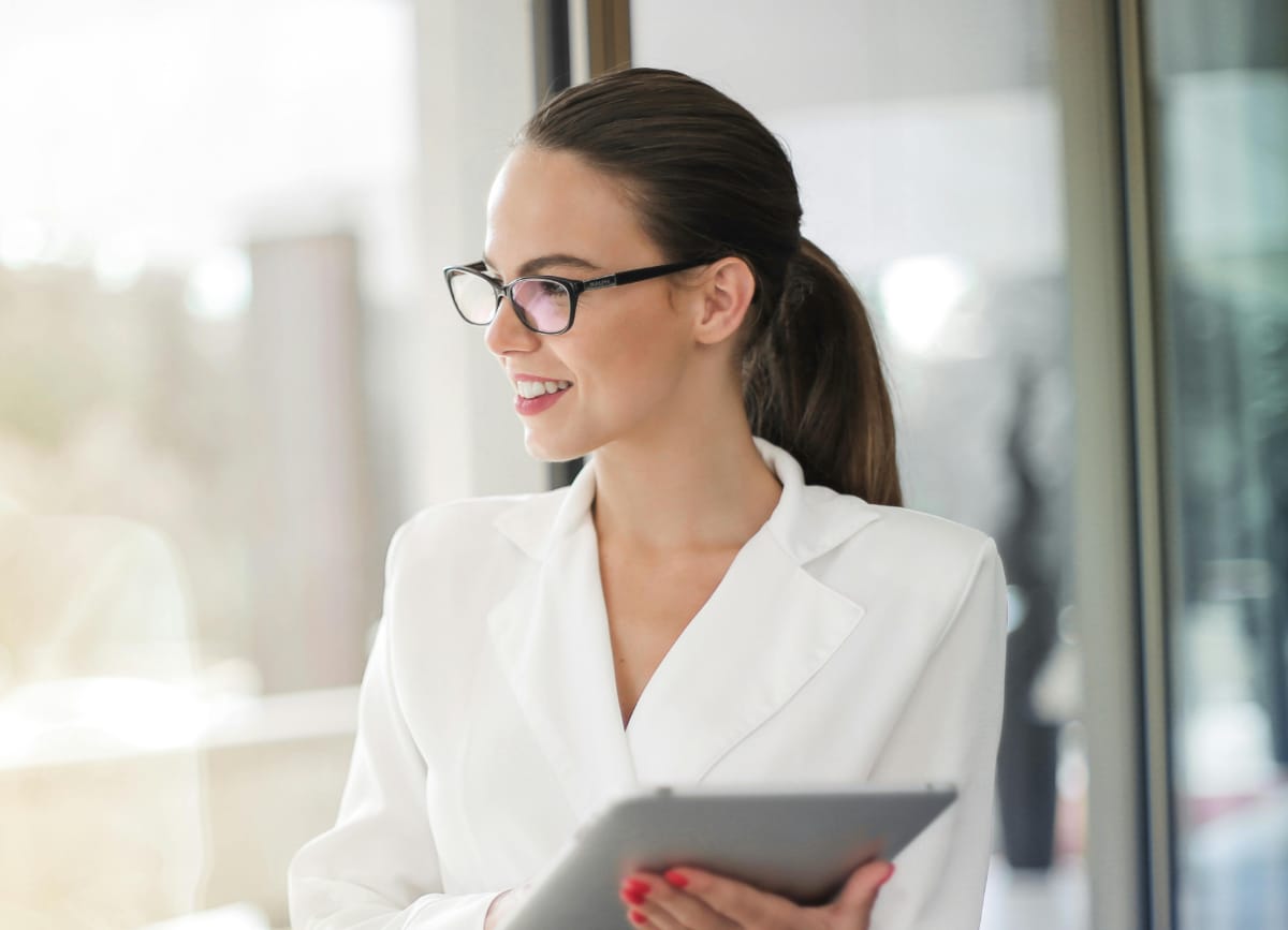 A photo of a professional businesswoman looking out of a skyscraper window holding a tablet in her hand